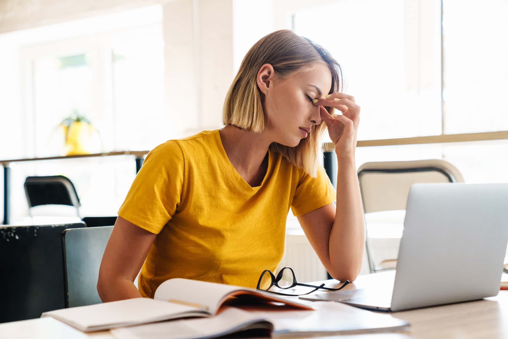 A woman is sitting at her desk and pressing her fingers to her nose bridge with her eyes closed.