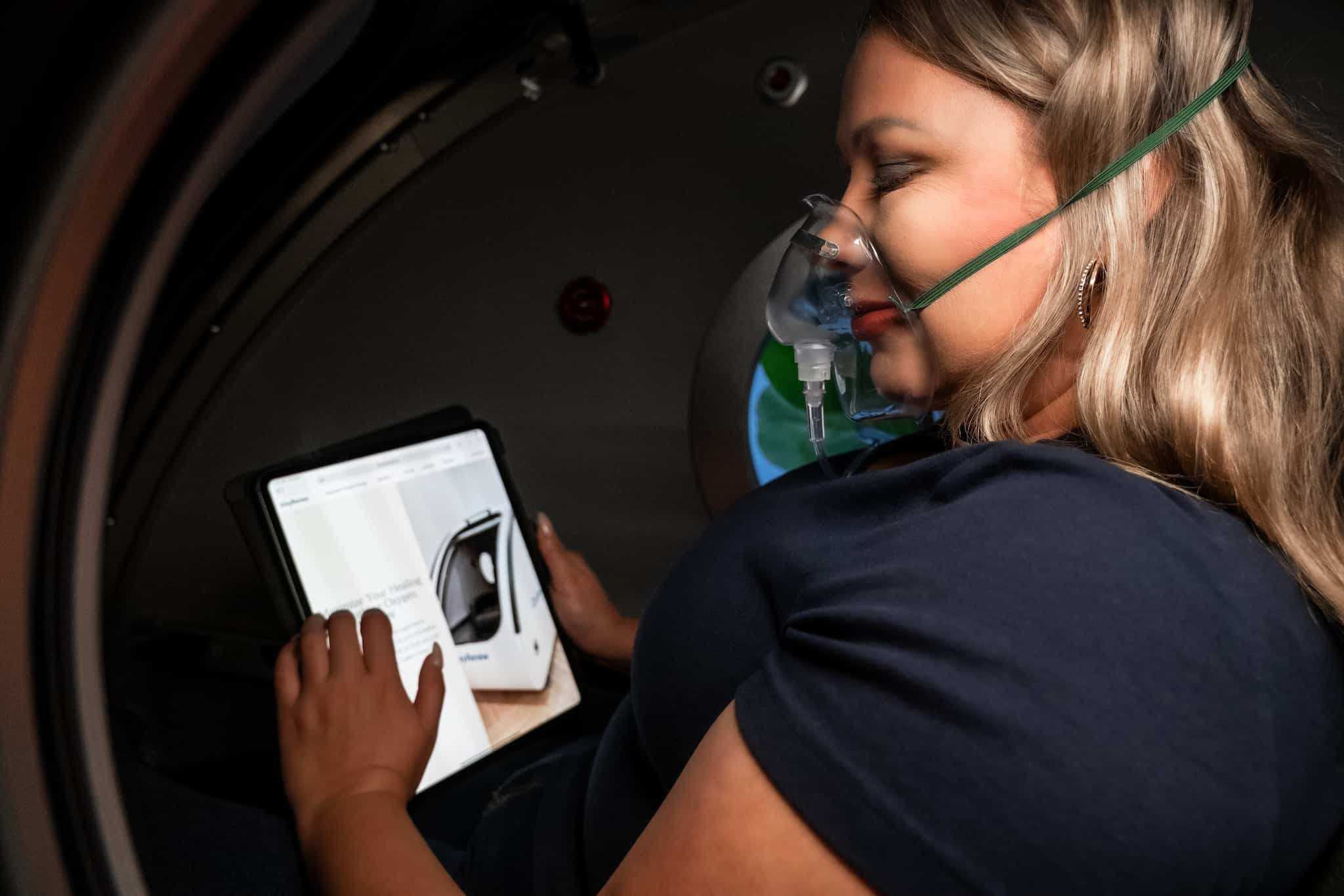 A woman is wearing an oxygen mask and using an iPad inside the OxyRenew hyperbaric oxygen therapy chamber.