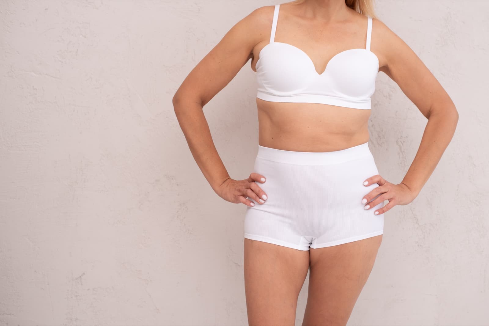 A woman in white underwear stands with her hands on her hips against a plain wall.